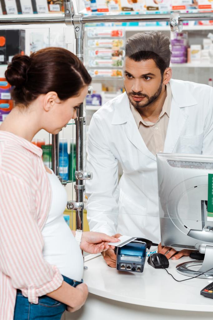 Farmacéutica mirando a una mujer embarazada pagando con un pase de pago en una farmacia
 - Foto, imagen