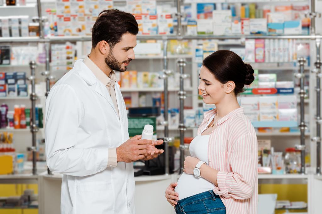 Vista lateral del farmacéutico mostrando frasco con pastillas a mujer embarazada sonriente en boticario
 - Foto, imagen