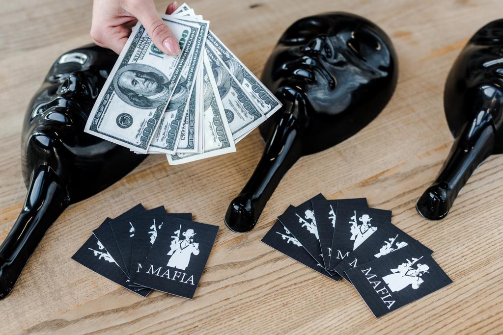 КИЕВ, УКРАИНА - 22 ноября 2019 года: обрезанный вид женщины, держащей долларовые банкноты рядом с игральными картами с мафиозными буквами и масками для вечеринок на столе
  - Фото, изображение