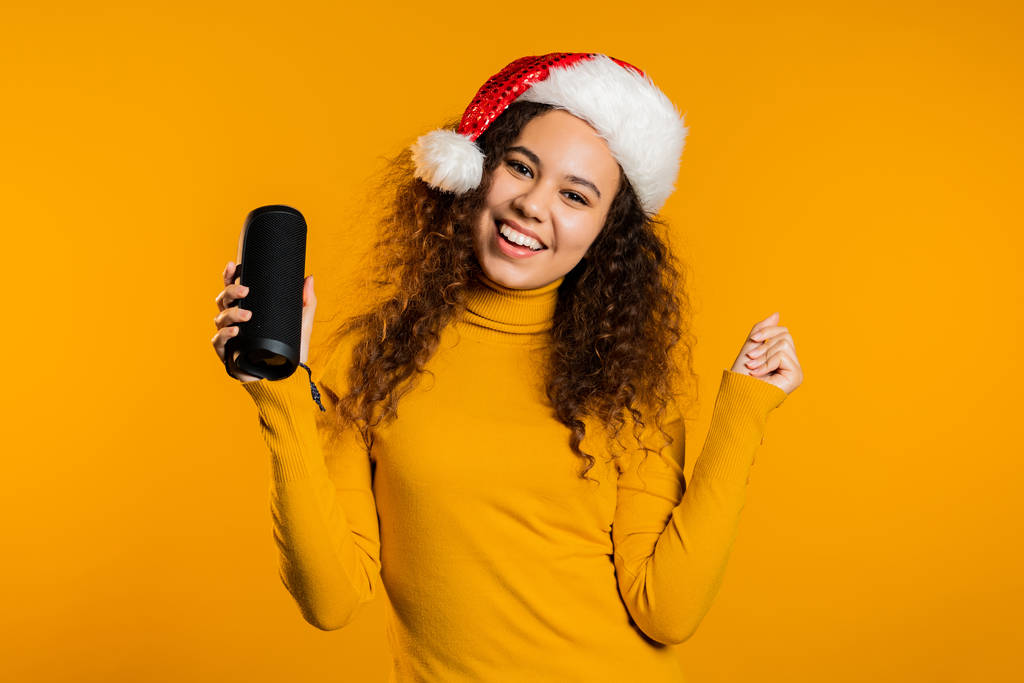 Young cute girl smiling and dancing with wireless portable speaker on yellow studio background. Woman in Santa hat. New year mood. - 写真・画像