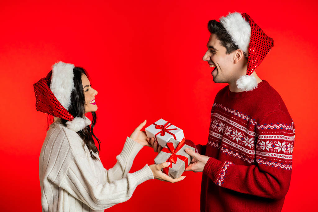 Jeune couple dans Noël chapeaux de Père Noël échanger des cadeaux sur fond de studio rouge. Amour, vacances, bonheur concept
. - Photo, image