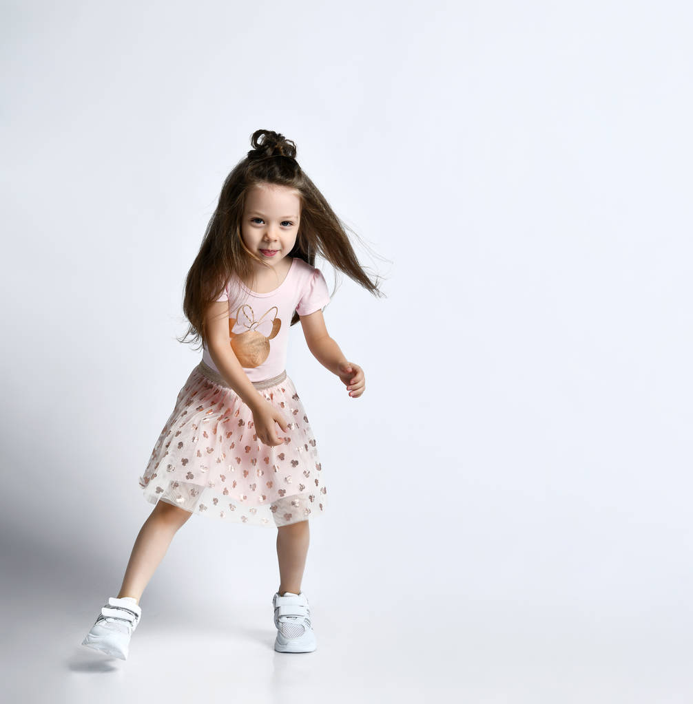 Маленька усміхнена мила дівчина в повсякденному літньому одязі та кросівки, що рухаються в дії на білому тлі стіни
 - Фото, зображення