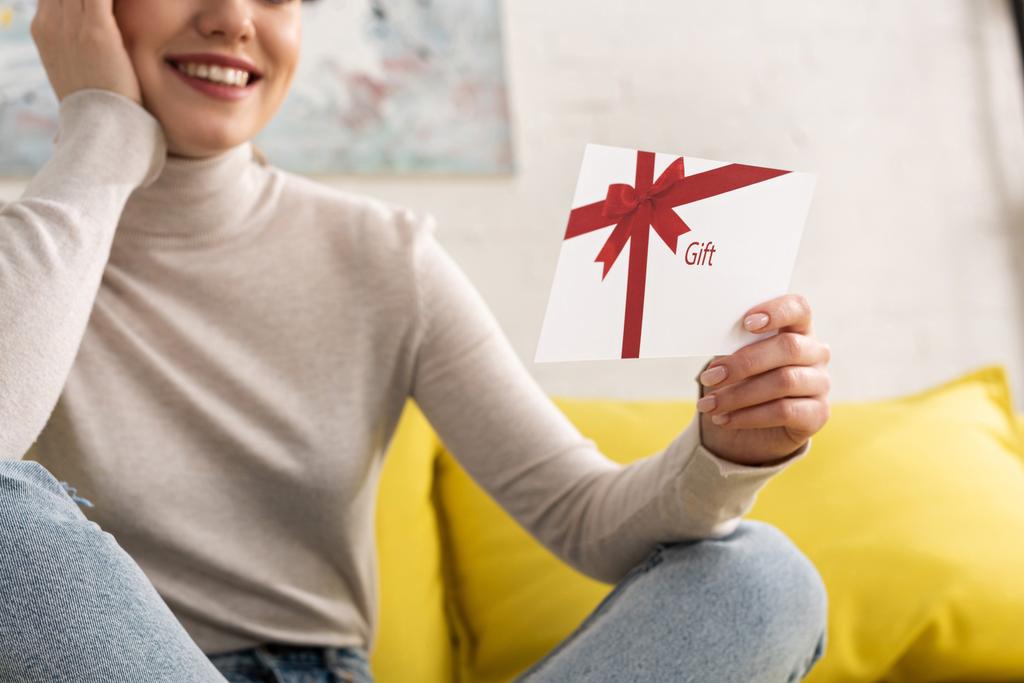 Обрезанный вид улыбающейся девушки с подарочной карточкой на диване
 - Фото, изображение
