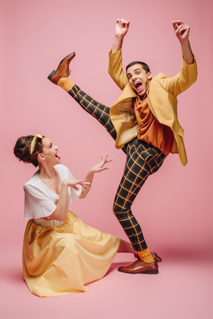 alegre chica sentado en piso y excitado hombre levantando la pierna mientras bailando boogie-woogie sobre rosa fondo
 - Foto, Imagen