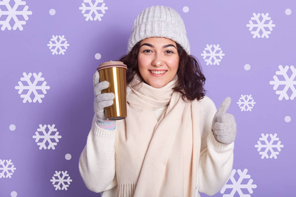 Usmívající se mladá žena drží termo hrnek s teplým nápojem a gestikuluje palcem nahoru, pózuje přes studiovou stěnu se sněhovou vločkou na pozadí, dáma v teplém zimním oblečení, žena je v dobré náladě. - Fotografie, Obrázek