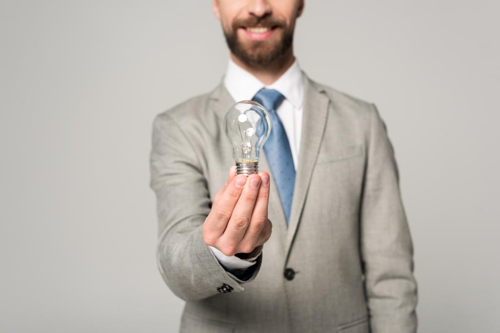 vista parcial de un empresario sonriente sosteniendo una bombilla aislada en gris
 - Foto, imagen