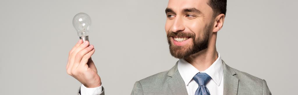 panoramiczne ujęcie uśmiechniętego biznesmena trzymającego żarówkę odizolowaną na szaro - Zdjęcie, obraz