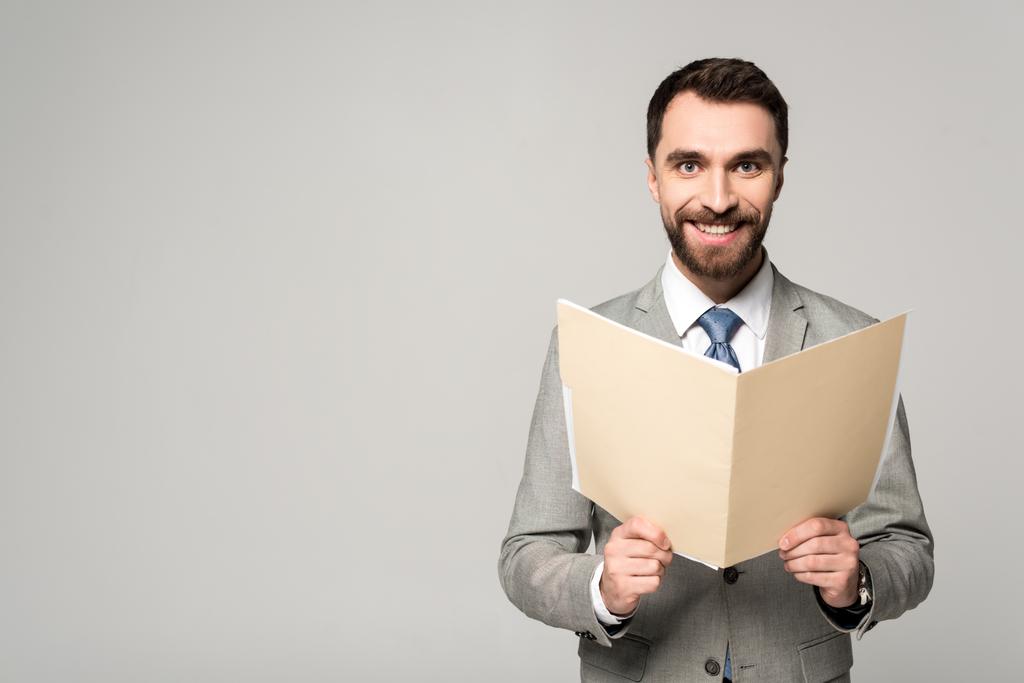 позитивный бизнесмен улыбается в камеру, держа бумажную папку изолированной на сером
 - Фото, изображение