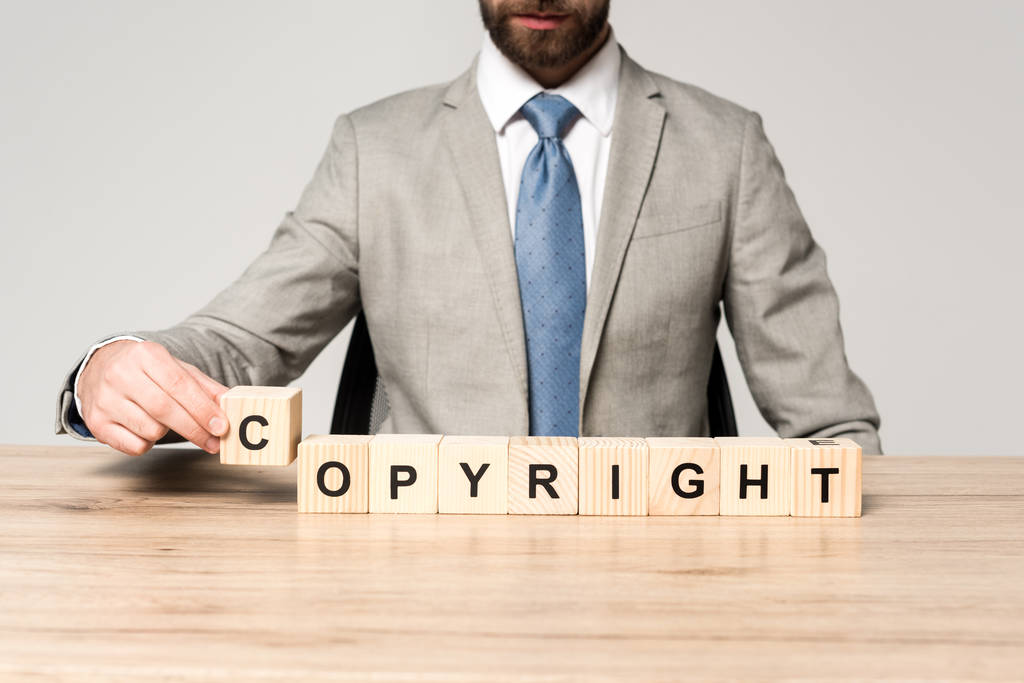 обрезанный взгляд бизнесмена, сидящего за столом рядом с деревянными кубиками со словом авторское право изолированы на серый
 - Фото, изображение