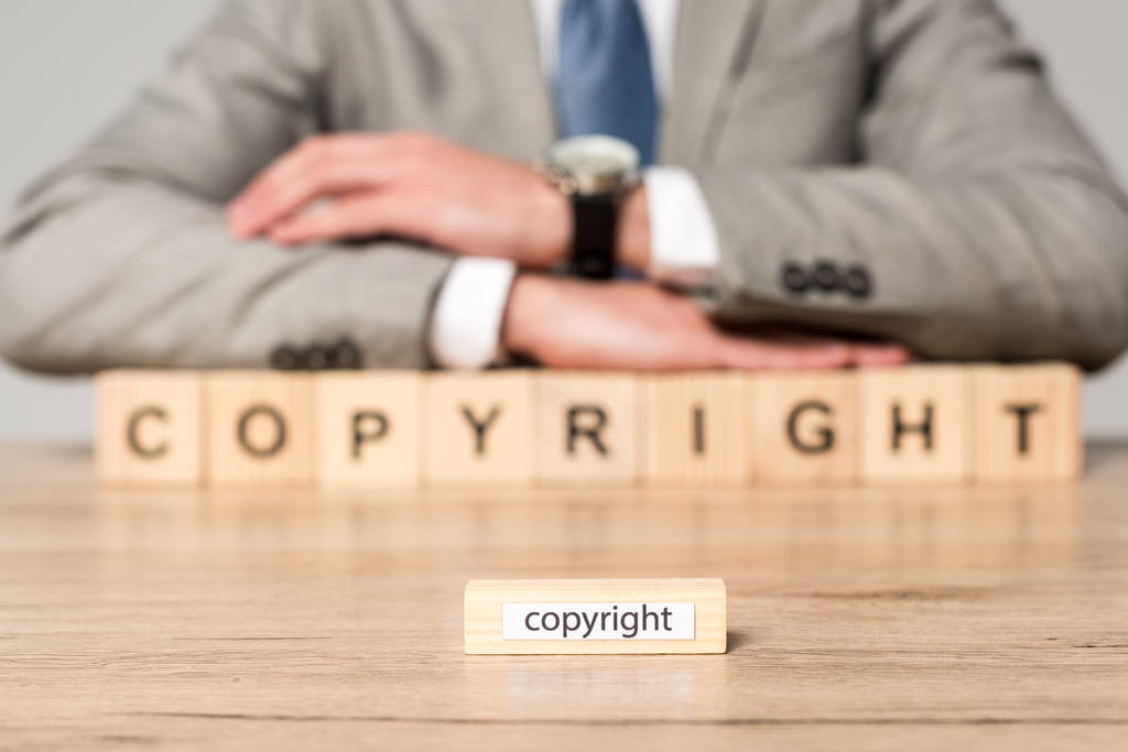 селективный фокус деревянного блока со словом авторского права, а бизнесмен сидит за столом рядом с деревянными кубиками с авторской надписью, изолированной на сером
 - Фото, изображение