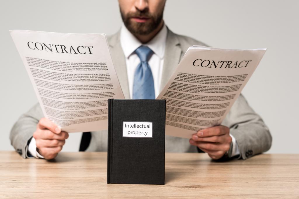 vista recortada del hombre de negocios leyendo contratos cerca de libro con título de propiedad intelectual aislado en gris
 - Foto, Imagen