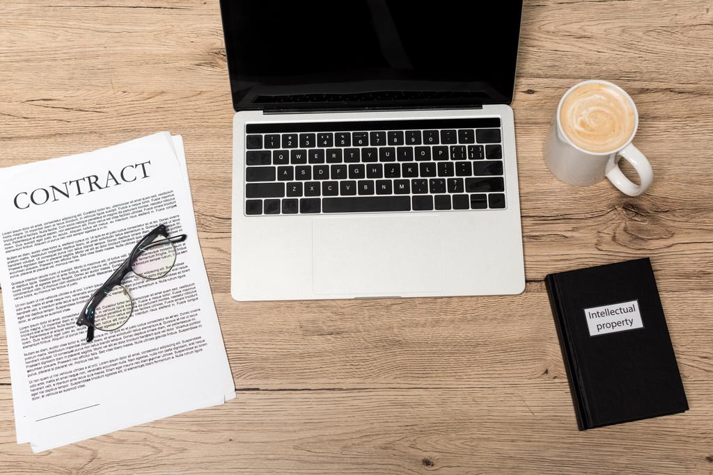 верхний вид ноутбука, контракт, очки, чашка кофе и книга интеллектуальной собственности на деревянном столе
 - Фото, изображение
