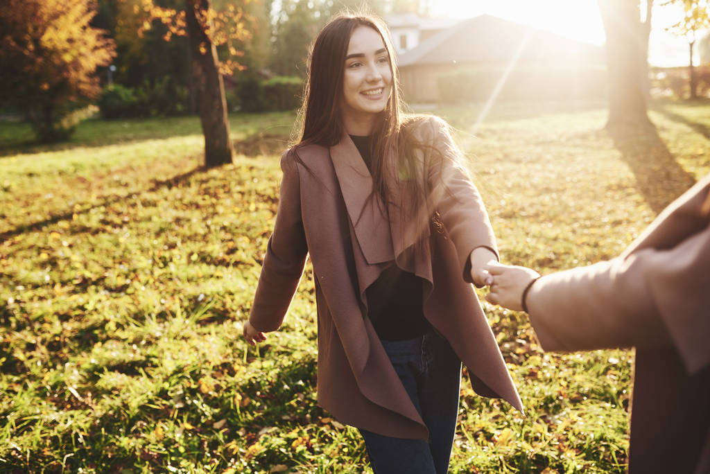 Young, smiling, pretty brunette girl is holding someone's hand while walking in autumn sunny park on blurry background. - Photo, Image