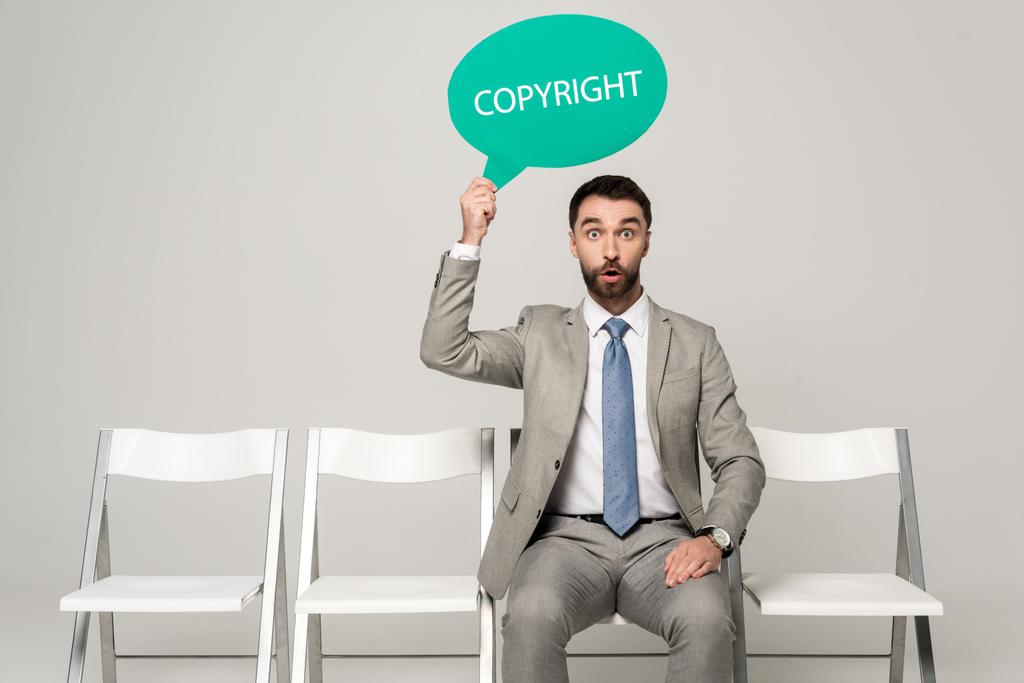 verrast zakenman houden gedachte zeepbel met woord copyright terwijl zitten op stoel op grijze achtergrond - Foto, afbeelding