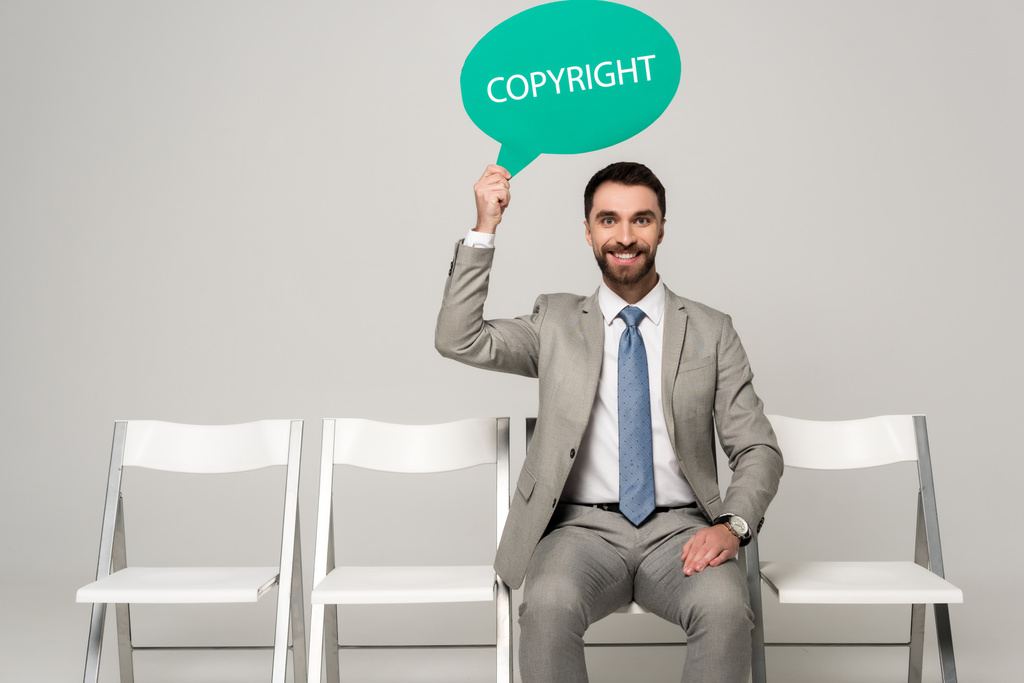 Lächelnder Geschäftsmann mit Gedankenblase mit Worturheberrecht, während er auf einem Stuhl auf grauem Hintergrund sitzt - Foto, Bild