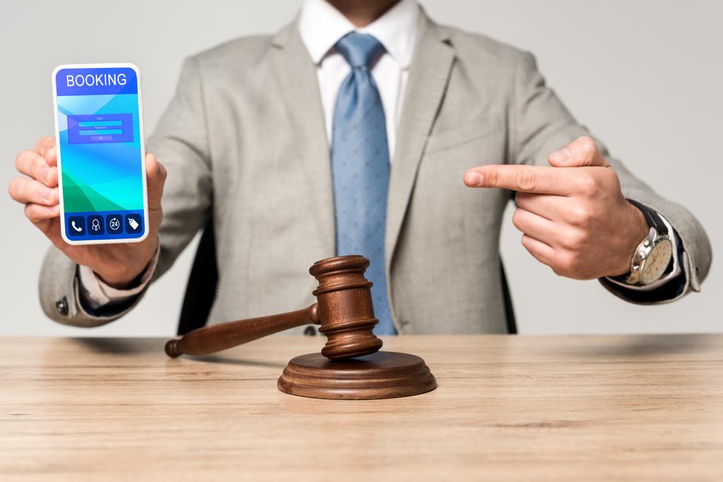 Ausgeschnittene Ansicht des Anwalts, der mit dem Finger auf Smartphone mit Buchungs-App zeigt, und Richtergabel auf dem Schreibtisch - Foto, Bild
