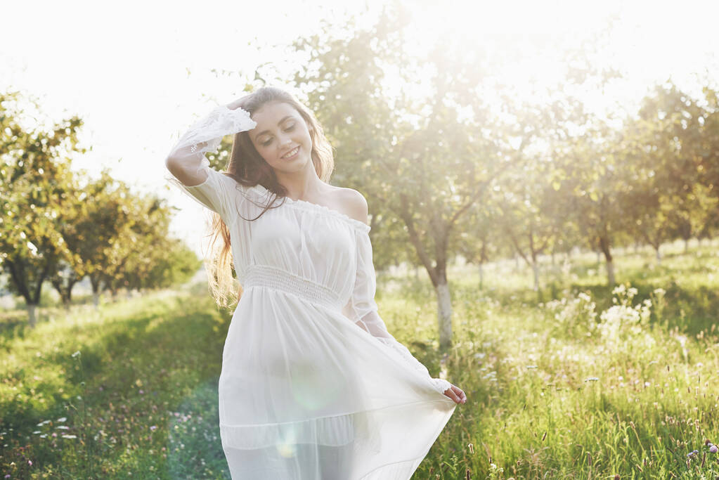 Nuori tyttö valkoisessa pitkässä mekossa kävelee puutarhassa. Kaunis auringonlasku puiden lehdissä
. - Valokuva, kuva