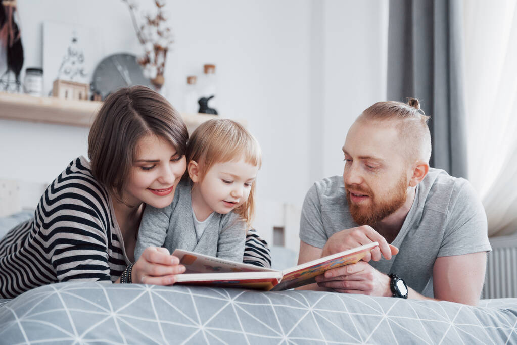 Isä, äiti ja pieni tytär lukemassa lastenkirjaa sohvalla olohuoneessa. Onnellinen iso perhe luki mielenkiintoisen kirjan juhlapäivänä. Vanhemmat rakastavat lapsiaan.
. - Valokuva, kuva