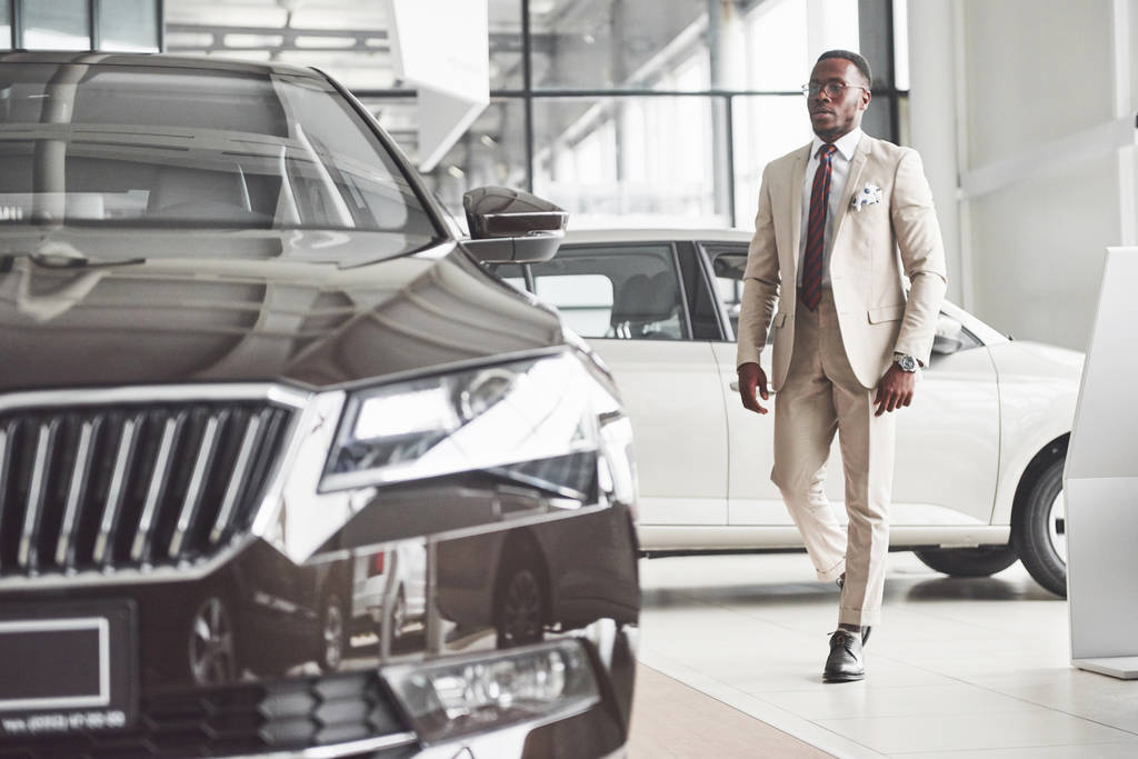 Jonge zwarte zakenman op autosalon achtergrond. Auto verkoop en verhuur concept. Rijke Afro-Amerikaanse man. - Foto, afbeelding