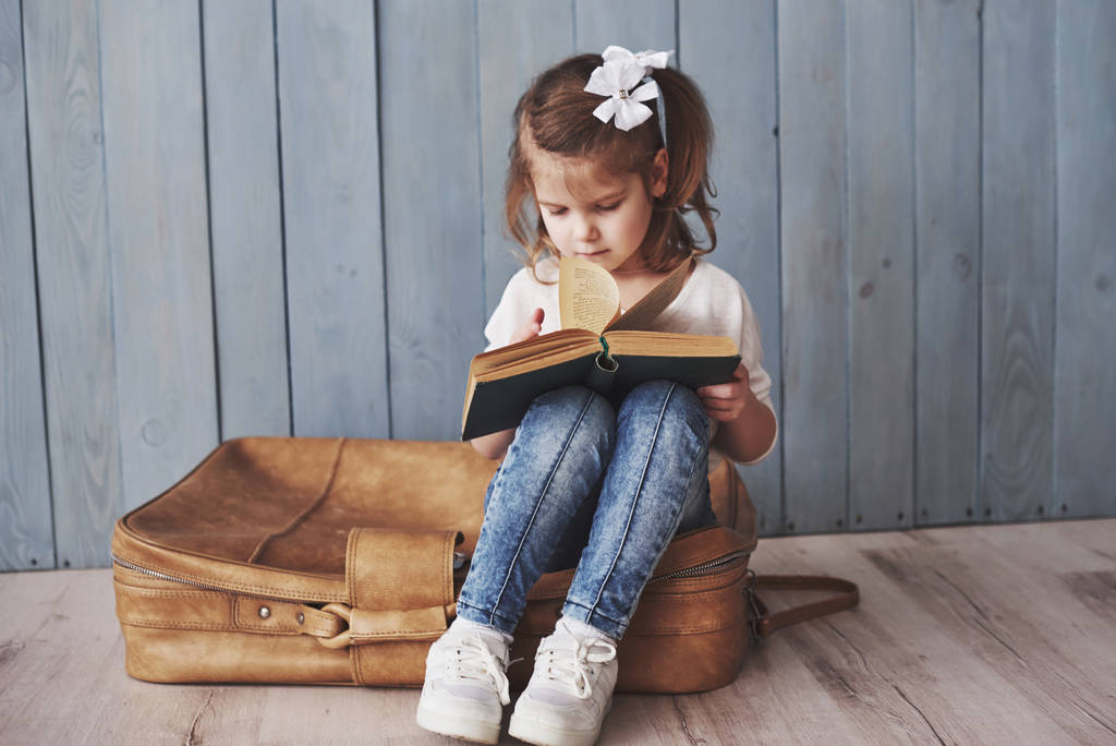 Готов к большому путешествию. Счастливая маленькая девочка читает интересную книгу с большим портфелем. Свобода и воображение
. - Фото, изображение