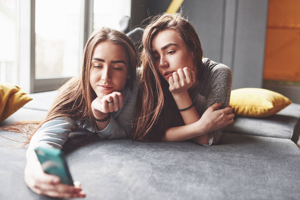 Dos hermanas gemelas sonrientes lindas sosteniendo el teléfono inteligente y haciendo selfie. Las niñas se acuestan en el sofá posando y alegría
. - Foto, Imagen