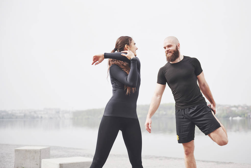 Couple de fitness s'étirant à l'extérieur dans le parc près de l'eau. Jeune homme barbu et femme faisant de l'exercice ensemble le matin
. - Photo, image