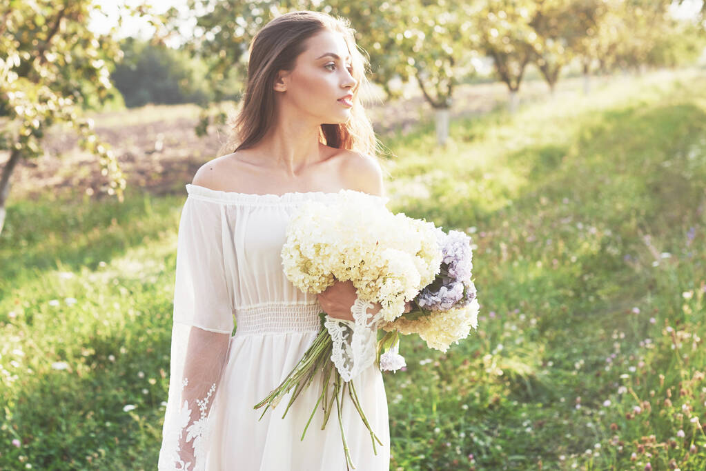 Красивая молодая девушка в белом светлом платье и букет летних цветов лежала прекрасный день в саду
. - Фото, изображение