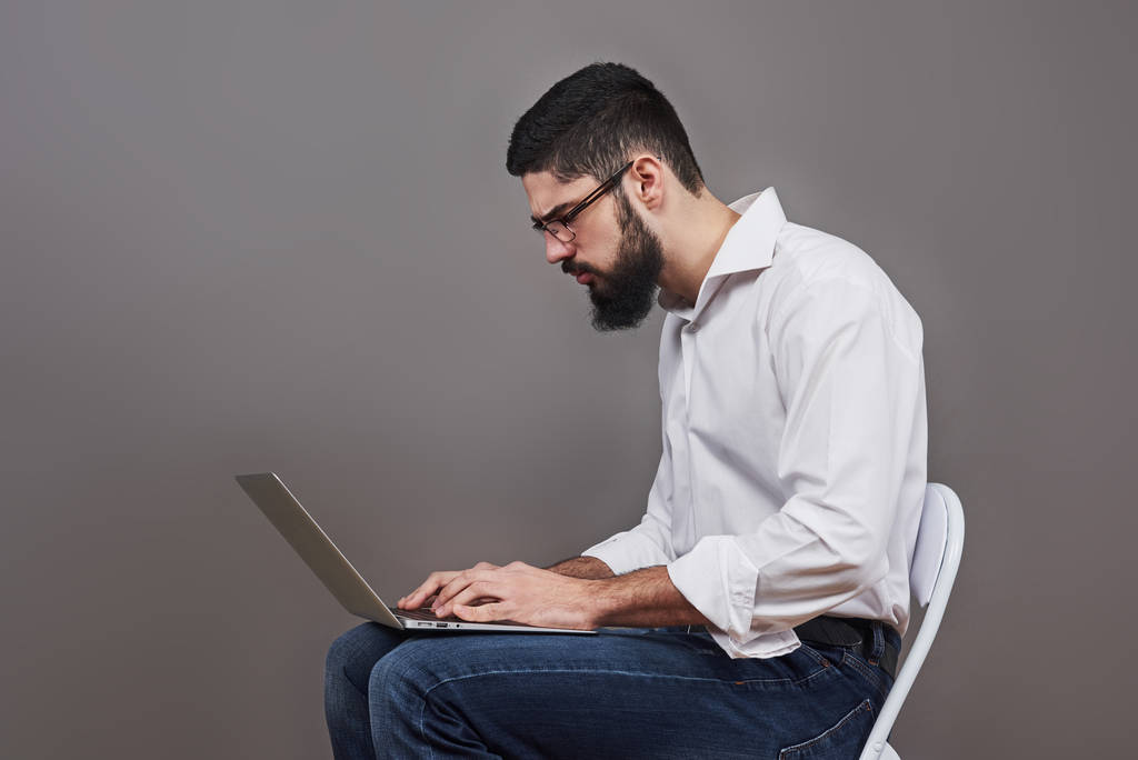Jóképű üzletember szemüveg és ruha laptop tartja a kezében, és írásban valamit. Oldalnézet. Elszigetelt szürke háttér. - Fotó, kép
