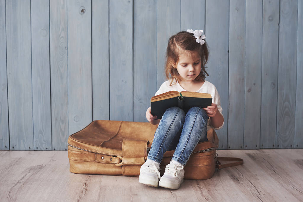Gotowy do wielkiej podróży. Szczęśliwa dziewczynka czytająca ciekawą książkę z dużą walizką i uśmiechnięta. Podróże, wolność i wyobraźnia. - Zdjęcie, obraz