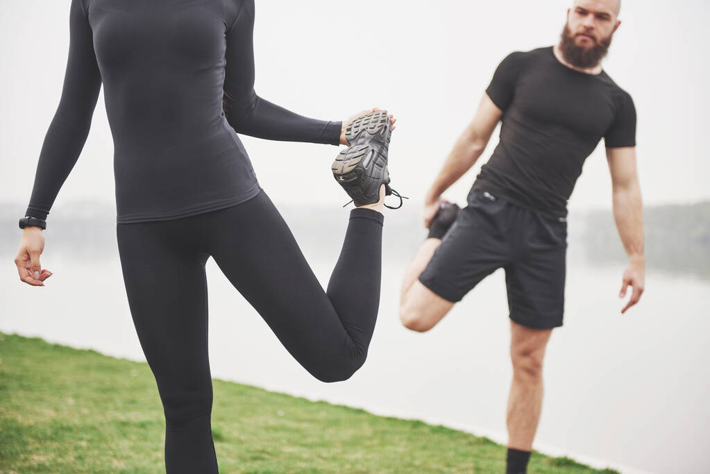 Fitness couple stretching outdoors in park near the water. Young bearded man and woman exercising together in morning. - Fotoğraf, Görsel