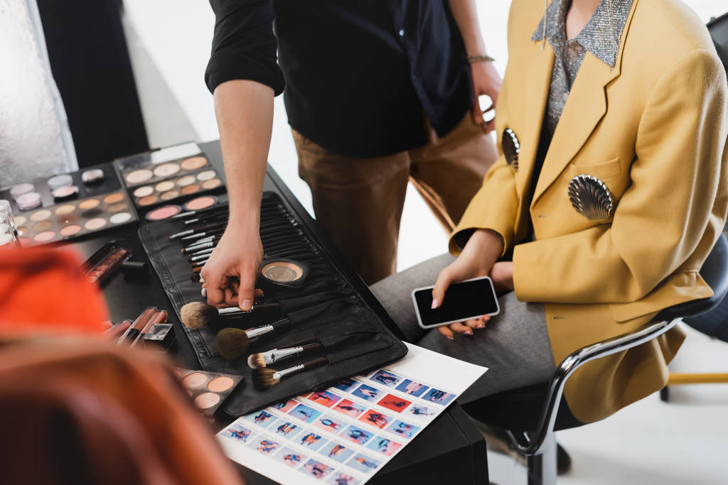 oříznutý pohled na make-up umělec s kosmetickým štětcem a modelem držící smartphone v zákulisí - Fotografie, Obrázek