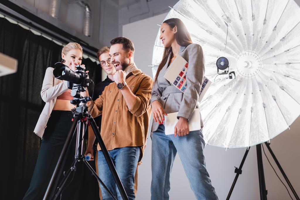 laag gezichtsveld van lachende fotograaf, model, stylist en producent kijkend naar digitale camera op backstage  - Foto, afbeelding