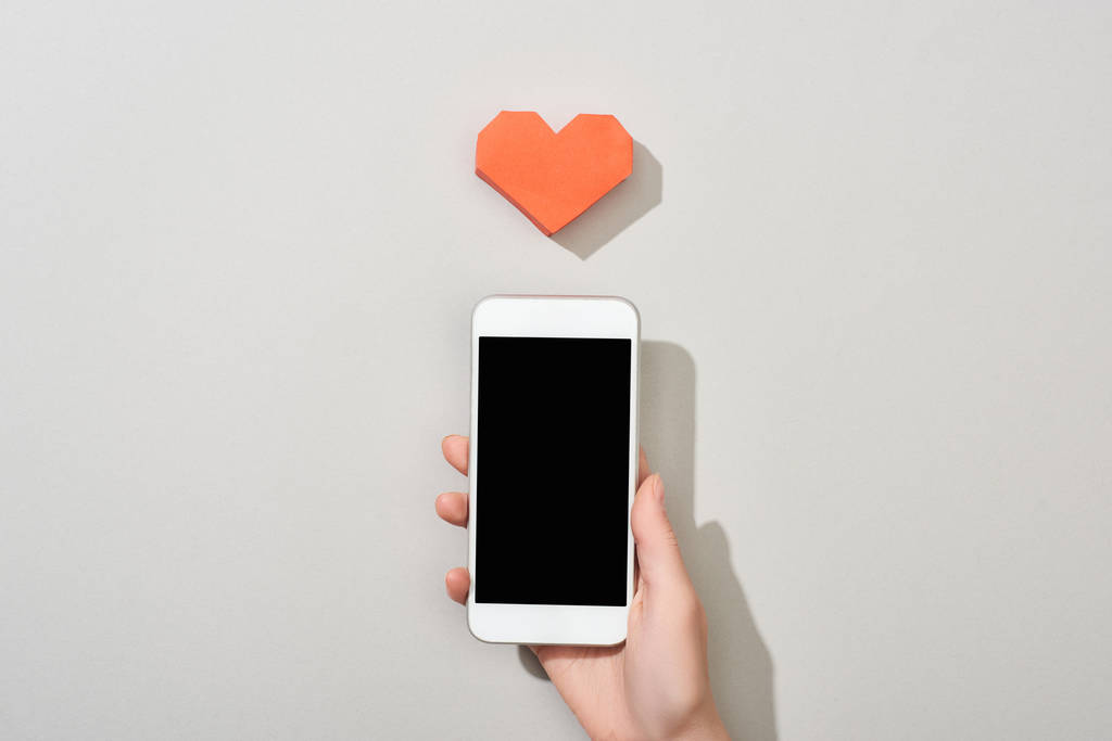 Visão superior da menina segurando smartphone com tela em branco por papel em forma de coração no fundo cinza
 - Foto, Imagem