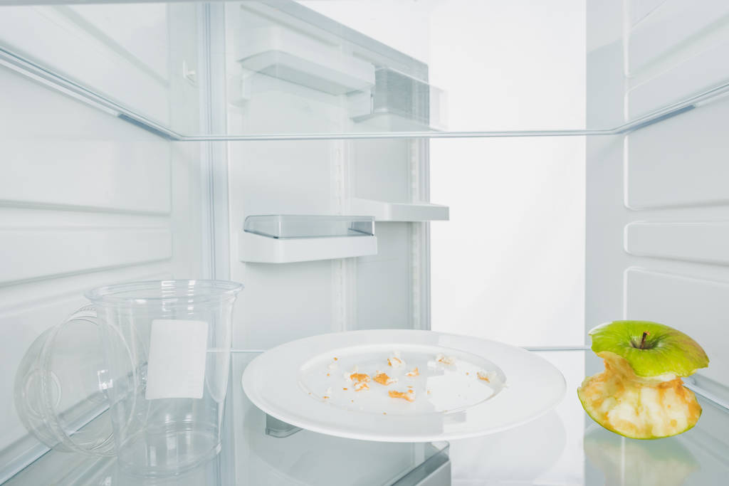 Plastikowy kubek z gryzionym jabłkiem i okruszkami na talerzu w lodówce z otwartymi drzwiami na białym - Zdjęcie, obraz