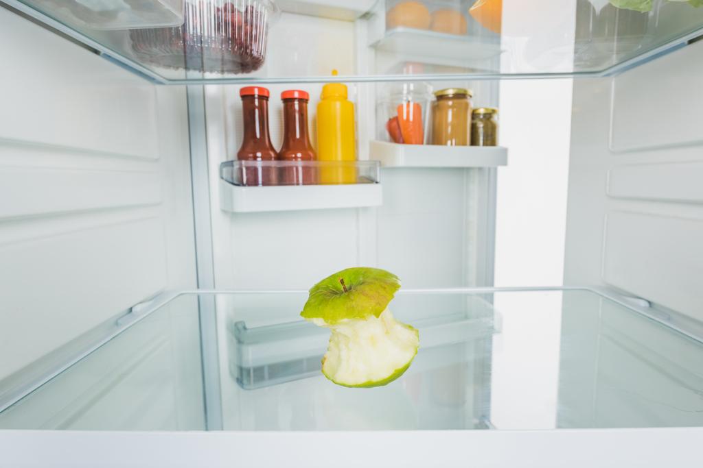 Επιλεκτική εστίαση του δαγκωμένου μήλου με σάλτσες σε ανοιχτή πόρτα ψυγείου που απομονώνονται σε λευκό - Φωτογραφία, εικόνα