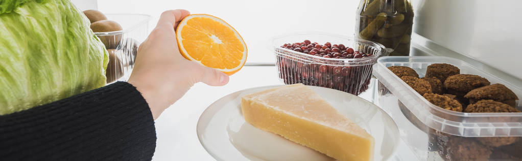 Καλλιεργημένη θέα της γυναίκας που παίρνει φέτα πορτοκαλιού από το ψυγείο με φαγητό απομονωμένο σε λευκό, πανοραμικό πλάνο - Φωτογραφία, εικόνα