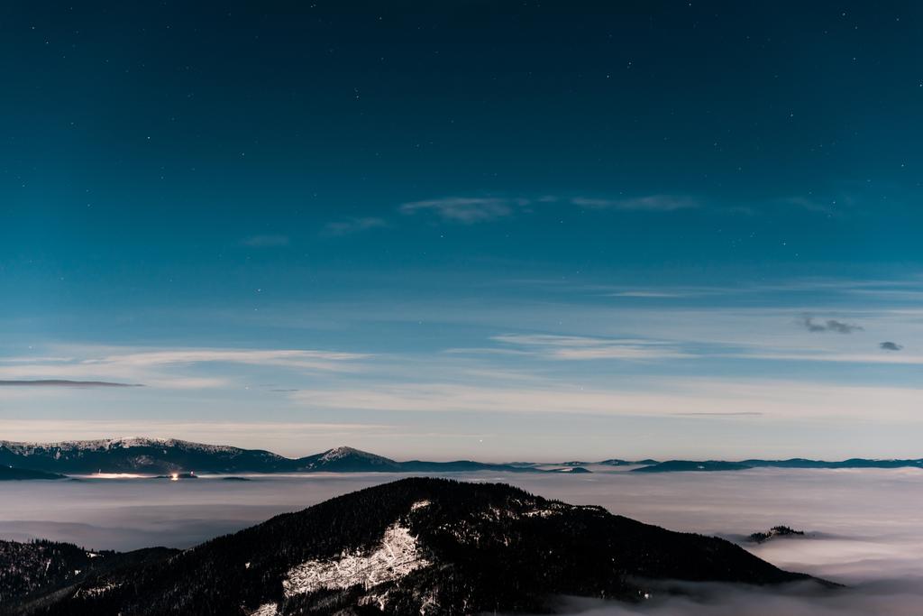 malebný pohled na zasněžené hory s borovicemi a bílé nadýchané mraky na tmavé obloze ve večerních hodinách - Fotografie, Obrázek