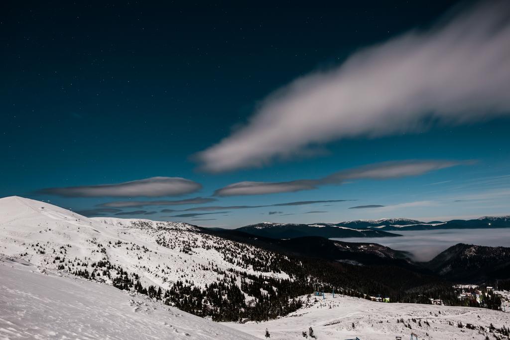 malowniczy widok na zaśnieżone góry z sosnami i białe puszyste chmury w ciemnym niebie wieczorem - Zdjęcie, obraz