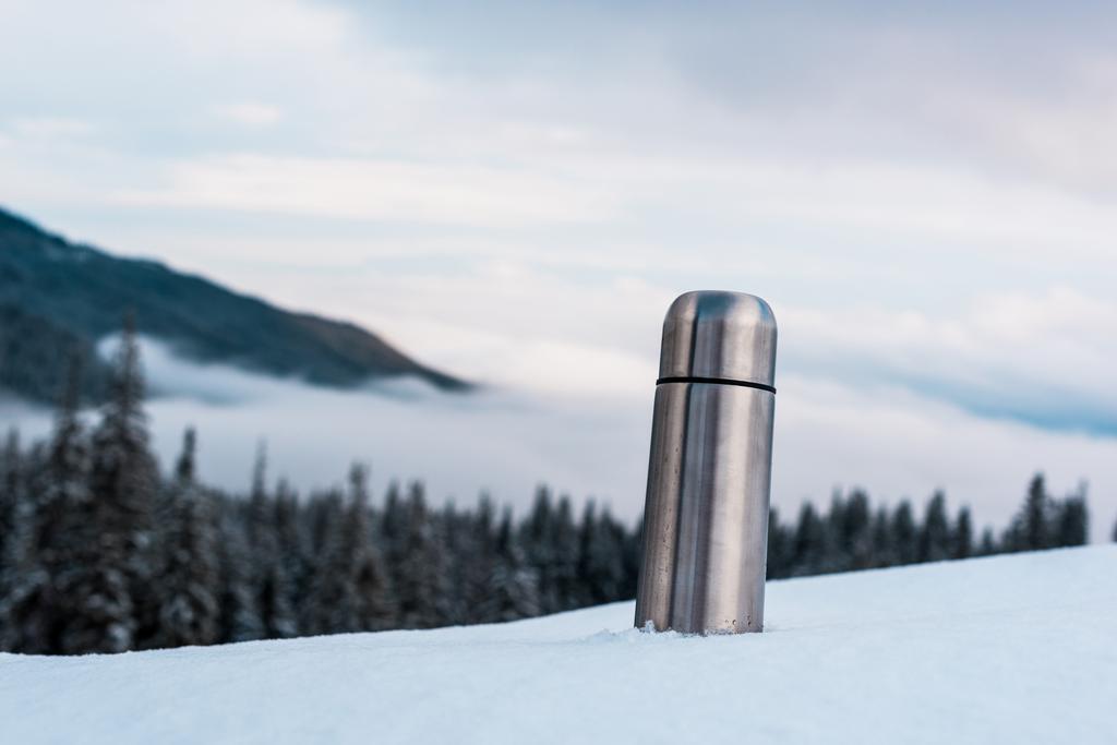 metallic vacuum flask in snowy mountains with pine trees and white fluffy clouds - Photo, Image