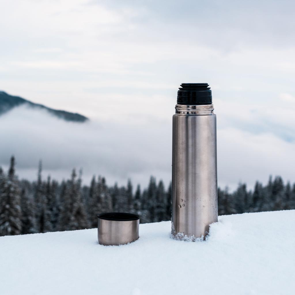 offene metallische Vakuumflasche in schneebedeckten Bergen mit Kiefern und weißen, flauschigen Wolken - Foto, Bild