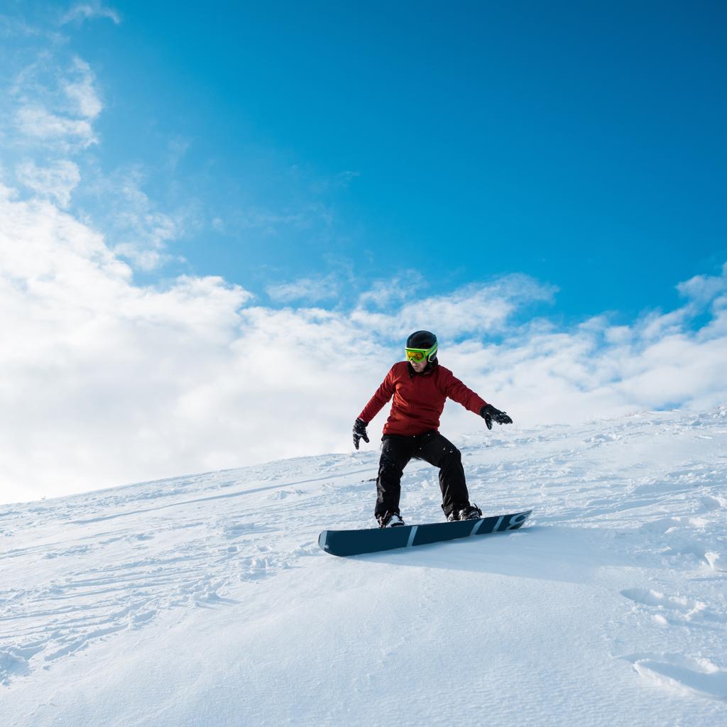 青い空に向かって斜面を滑り降りるヘルメットのスノーボーダー  - 写真・画像