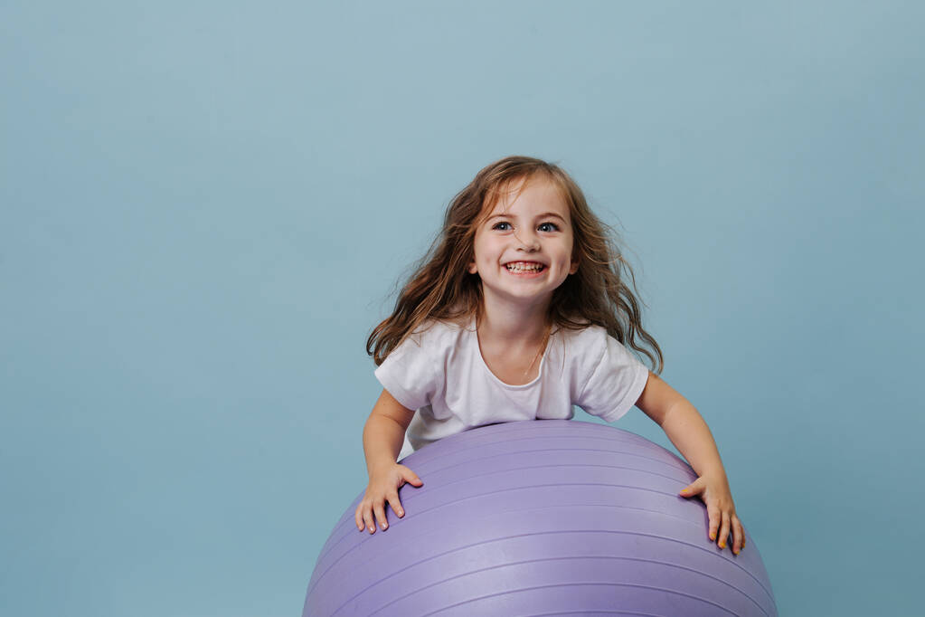 blue-eyed curly toddler girl laughs playing on lilac fitness ball. - Photo, Image