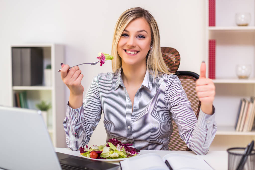 Gyönyörű üzletasszony élvezi eszik saláta és mutatja hüvelykujját fel, miközben dolgozik az irodájában. - Fotó, kép