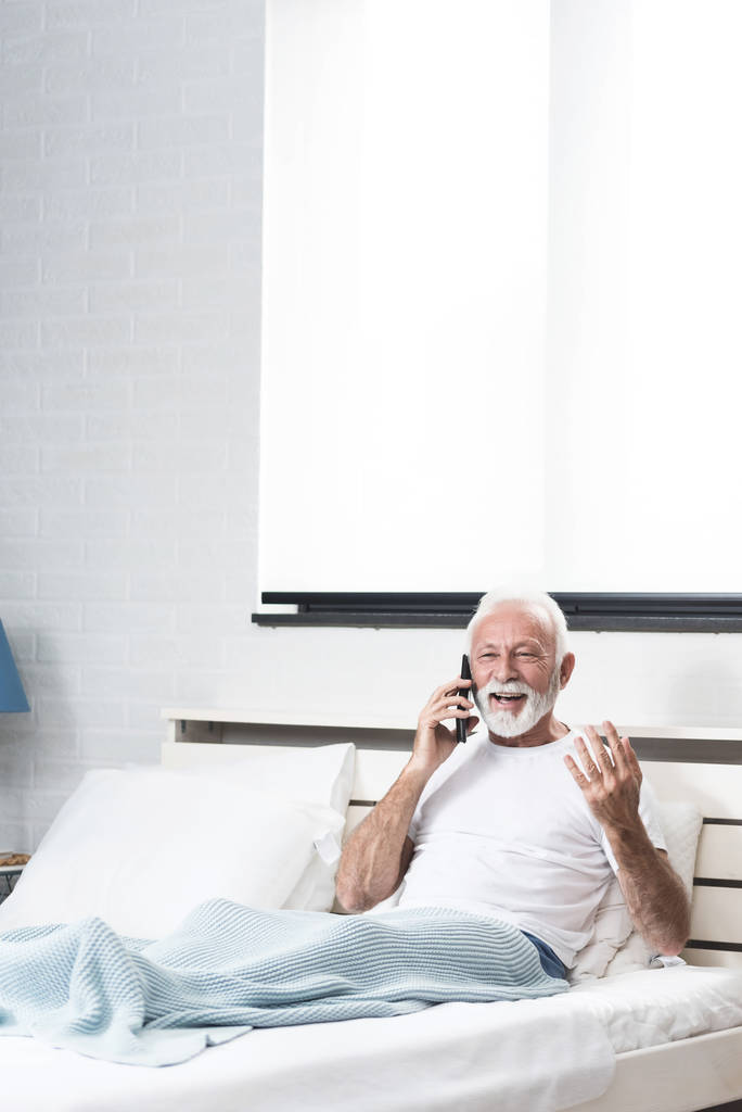 Idősebb férfi kap pozitív híreket telefonon keresztül az ágyban a szobájában. Pizsamában beszélt telefonon, miután nagy mosollyal ébredt. Meglepett arc. Helykép másolása - Fotó, kép