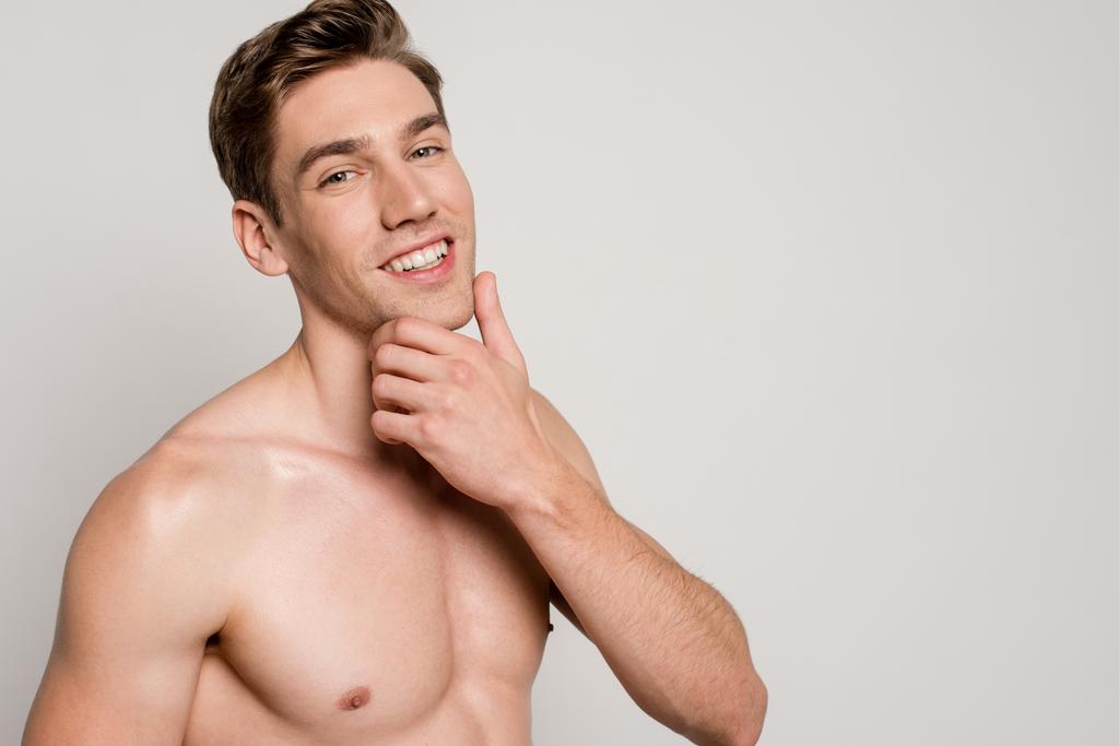 улыбающийся сексуальный мужчина с мускулистым туловищем, касающимся подбородка, изолированный на сером
 - Фото, изображение