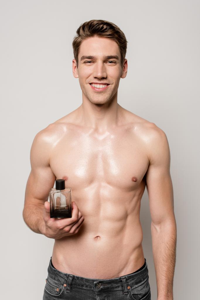 usmívající se sexy muž s svalnatým trupem drží parfém láhev izolované na šedé - Fotografie, Obrázek