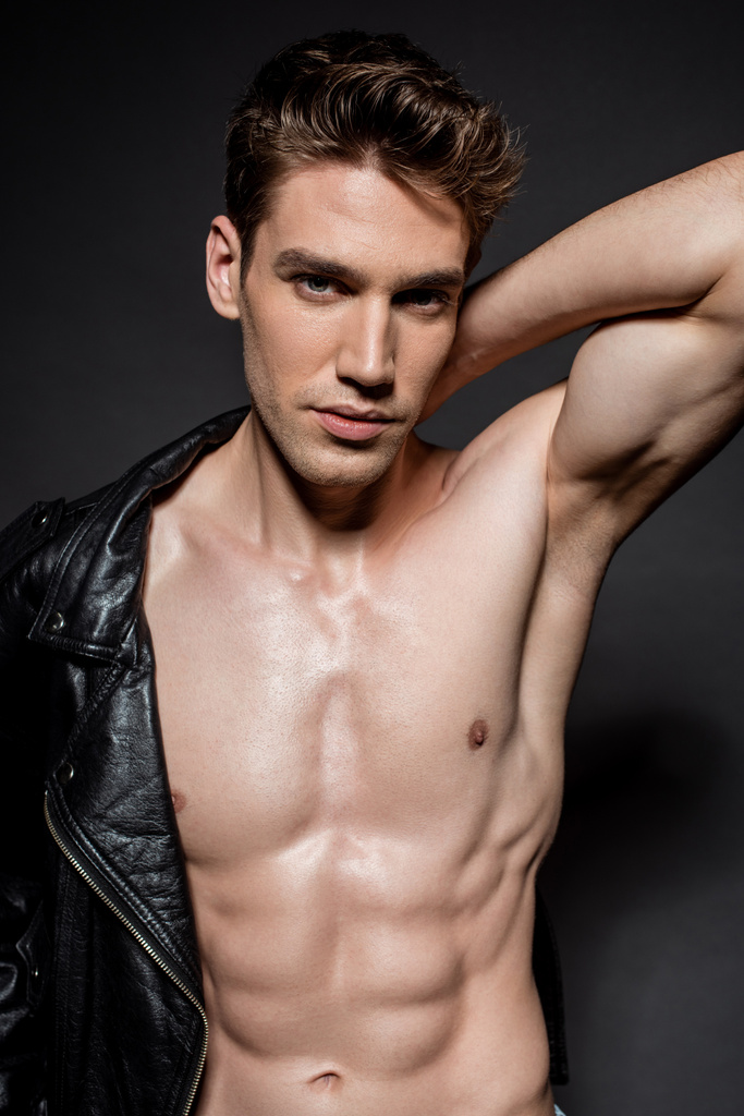sexy young man with muscular torso putting on biker jacket on black background - Photo, Image