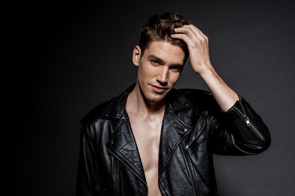 handsome sexy young man in biker jacket touching hair on black background - Photo, Image