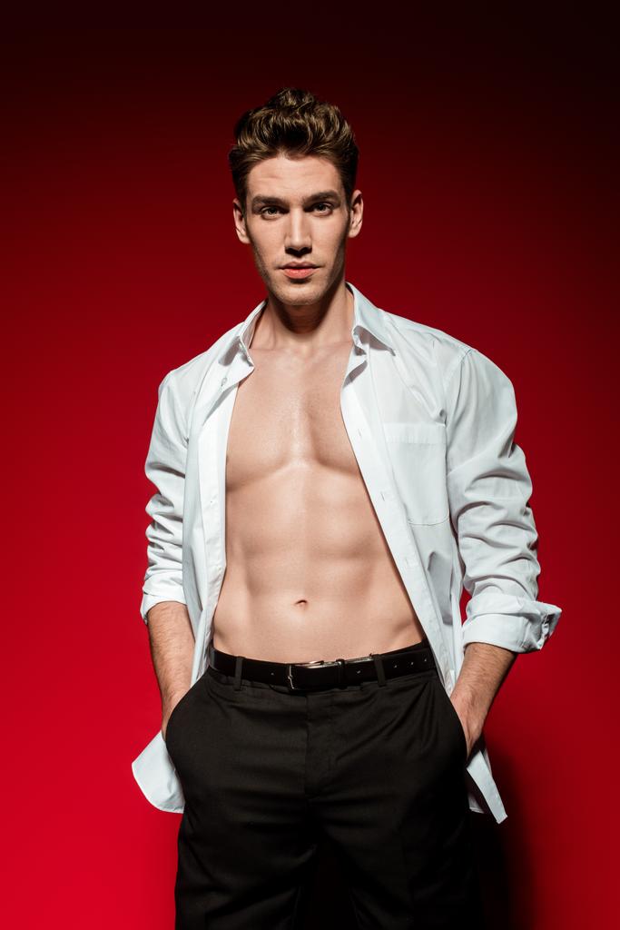 sexy młody elegancki mężczyzna w rozpięta koszula z muskularny goły tułów i ręce w kieszeniach na czerwonym tle - Zdjęcie, obraz