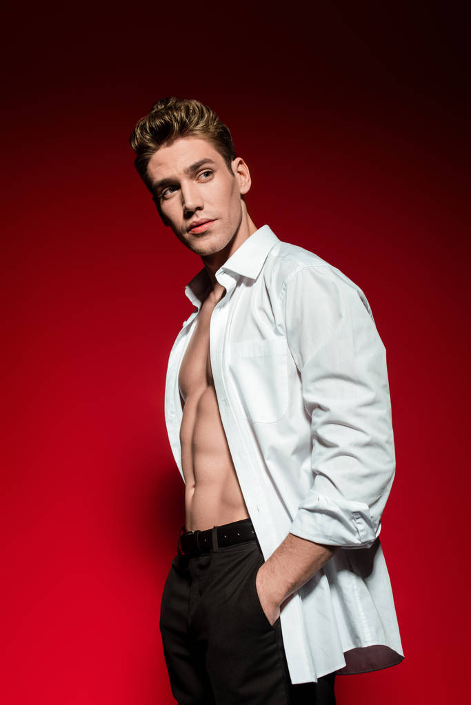 сексуальный молодой элегантный мужчина в расстегнутой рубашке с мускулистым туловищем и рукой в кармане, глядя в сторону на красный фон
 - Фото, изображение
